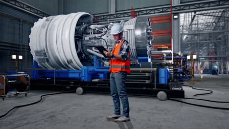 full body side view of asian male engineer with safety helmet working on a tablet while standing with airplane engine maintenance conducted, analytics checking the turbine