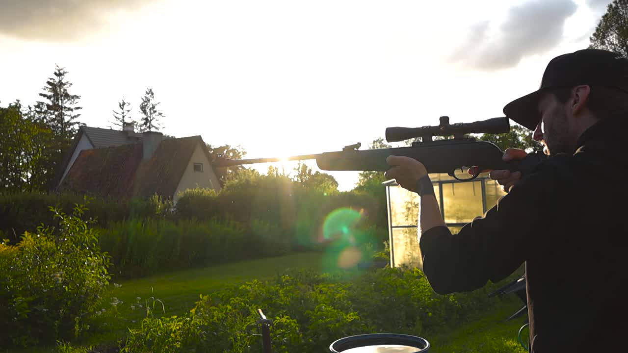 Man lifting up and shooting a gun rifle firearm during a sunset day in a garden in slow motion with lense and light flare. Man has black clothing and aims the gun by watching through scope telescope