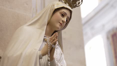 estatua serena de la virgen maría, manos juntas en oración, adornada con una corona y sosteniendo cuentas de rosario, simbolizando devoción y fe