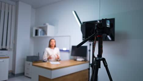 female podcast host sitting at desk speaking emotionally to camera. day light lamp illuminates the speaker. blurred backdrop.