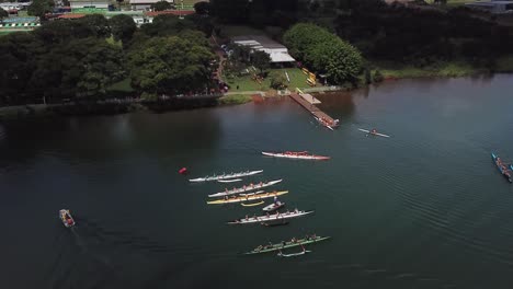 aerial descending view towards colourful group of long athletes row boats on lake water