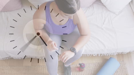 digital clock ticking against overhead view of caucasian woman using smartwatch at home