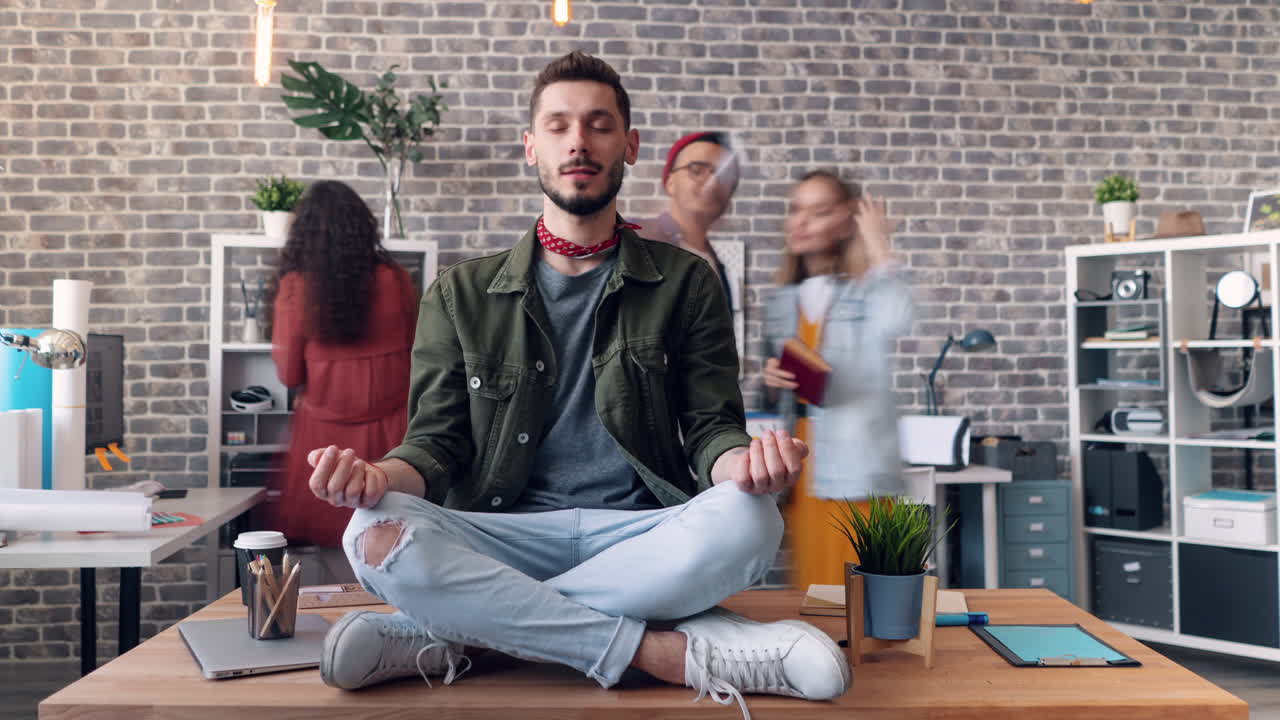 Man meditating at his desk in an office, while colleagues are working around him