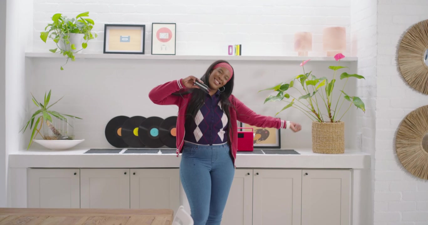 African American woman in headband dancing, holding phone playing music near red speaker in kitchen