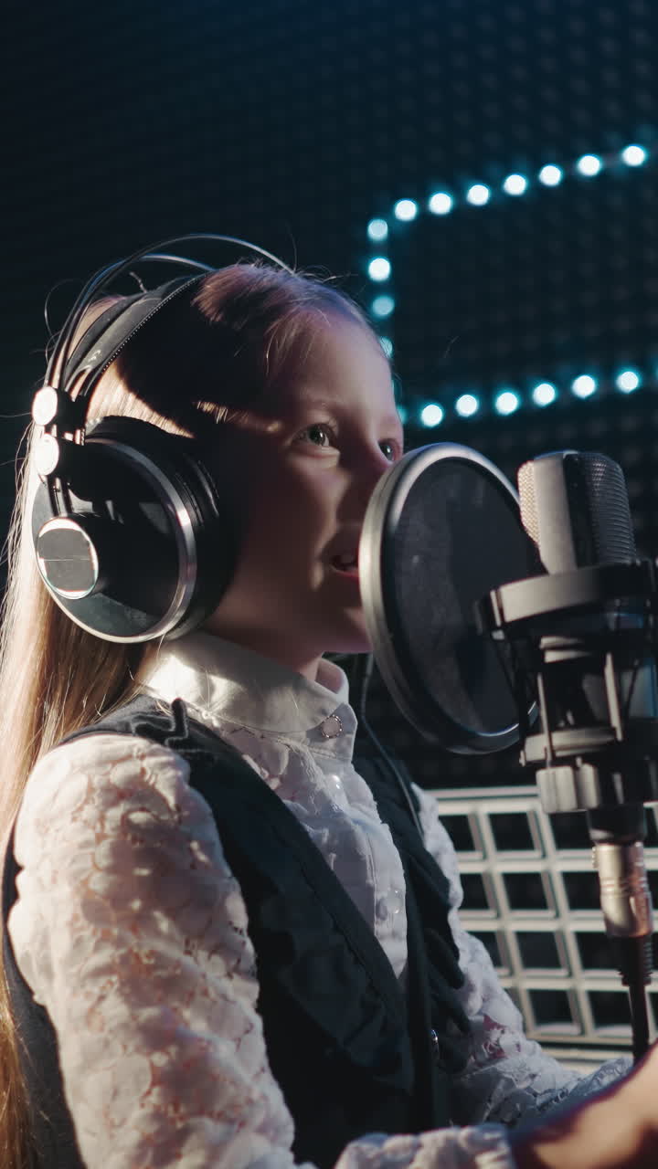 Girl holds on microphone stand singing. Determined small girl tries singing notes correctly in recording studio. Recording of song by young artist