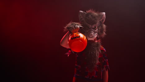 studio shot of child dressed up in werewolf monster costume trick or treating at halloween with red smoke background lighting holding pumpkin shaped jack o'lantern bucket 1