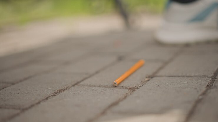 Small object lands on pavement quietly. Brief slip causes pencil to land unexpectedly on path. An orange pencil accidentally drops onto brick surface near sneaker in tranquil park
