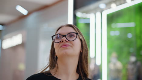 primer plano de un estudiante con gafas y camiseta negra caminando por un centro comercial moderno, mirando a su alrededor con curiosidad y aprecio, con luces de neón brillantes y exhibiciones de tiendas elegantes