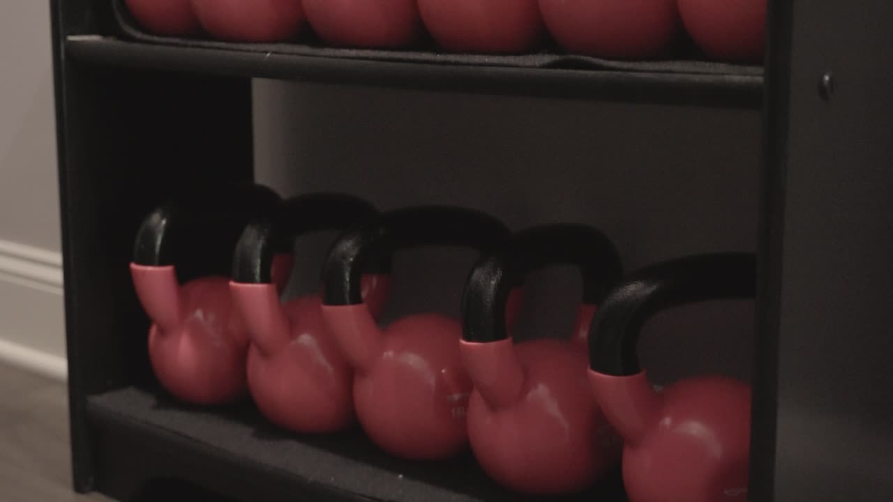 In a professional gym setting, a rack displaying gym equipment such as dumbbells and kettlebells is present
