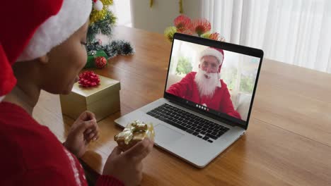 Afroamerikanische-Frau-Mit-Weihnachtsmannmütze-Nutzt-Laptop-Für-Weihnachtsvideoanruf,-Mit-Weihnachtsmann-Auf-Dem-Bildschirm