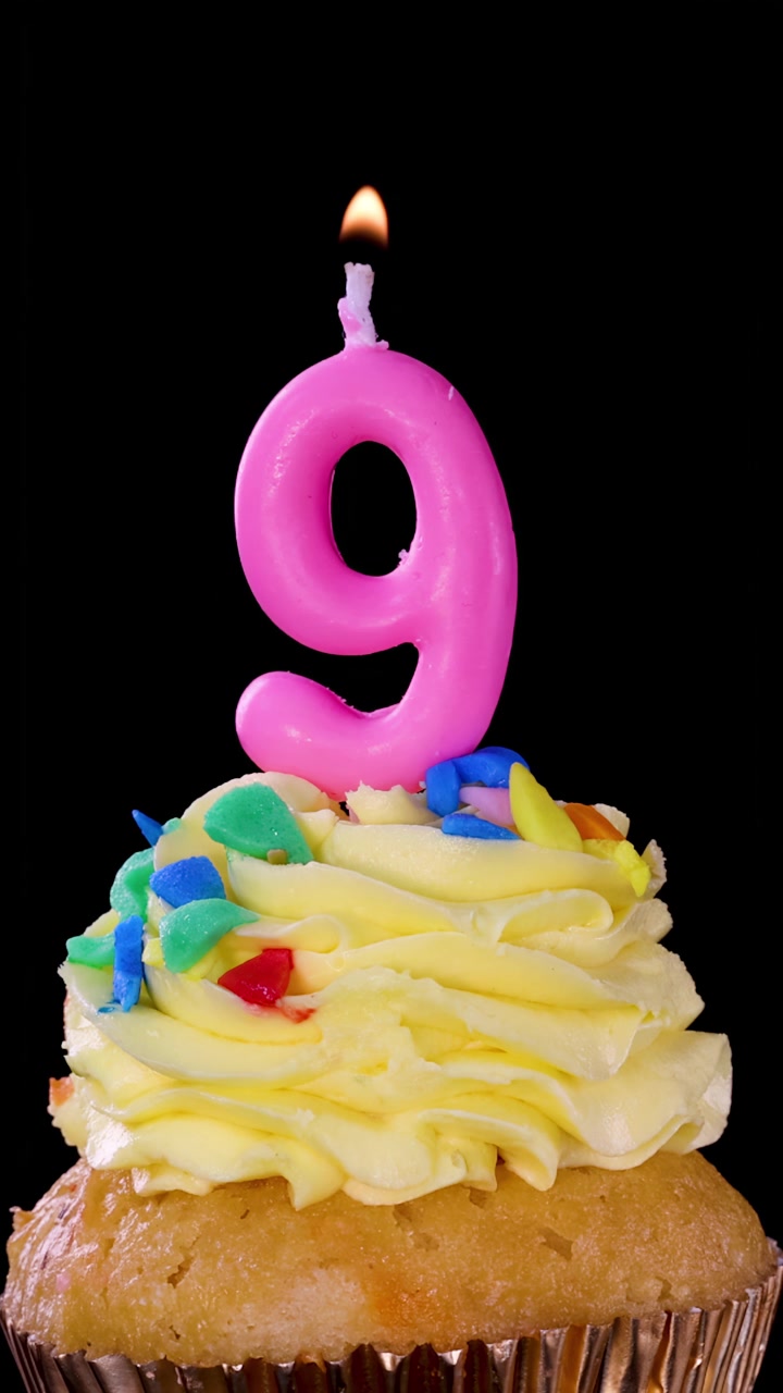 Una mano accende una candela rosa a forma di numero nove su un cupcake colorato con un accendino, sfondo nero, illuminazione brillante.