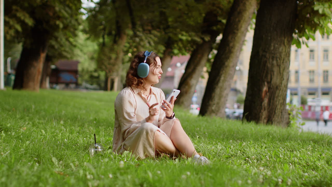 Happy young woman in wireless headphones choosing listening music in smartphone dancing outdoors