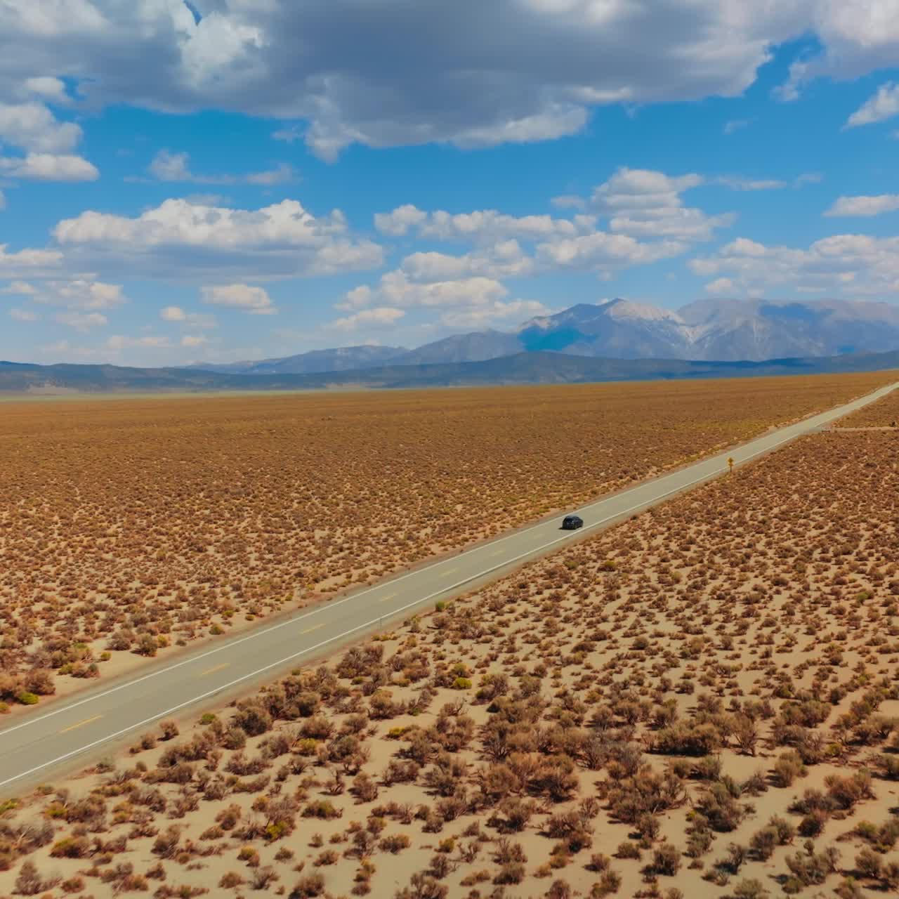 Lonely black car riding by the dry road from California to Nevada. Amazing blue skies with cotton clouds over mountains at backdrop