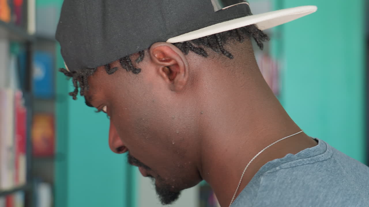 Close up side view of young man in casual shirt and backward cap standing in library, focused on books arranged neatly on shelf, appearing deep in thought while scanning titles