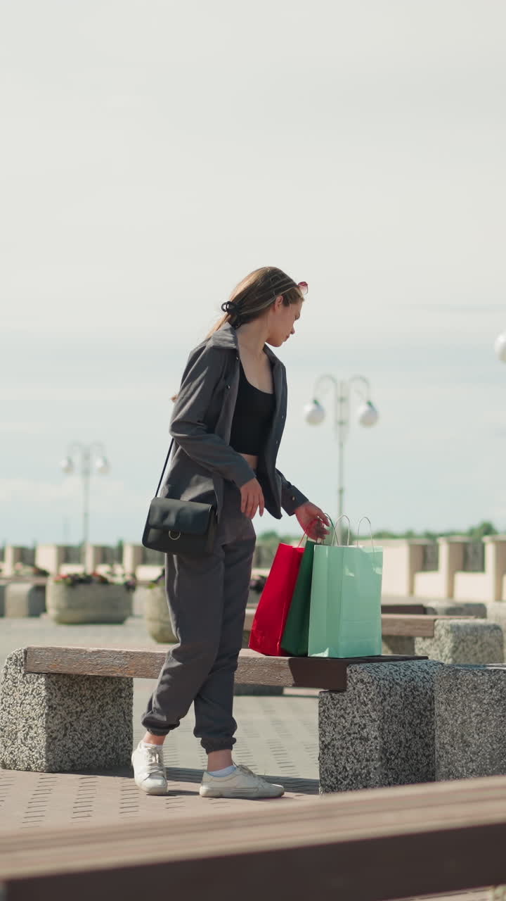 una joven con ropa gris se sienta al aire libre, dejando caer su bolsa de compras cerca, en el fondo, una madre e hija se toman de la mano mientras caminan, vista borrosa de la gente en la distancia