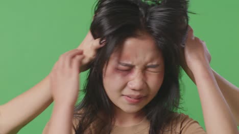 close up of young asian woman victim of violence with bruise on face abused by man's hands pulling hair on green screen background in the studio