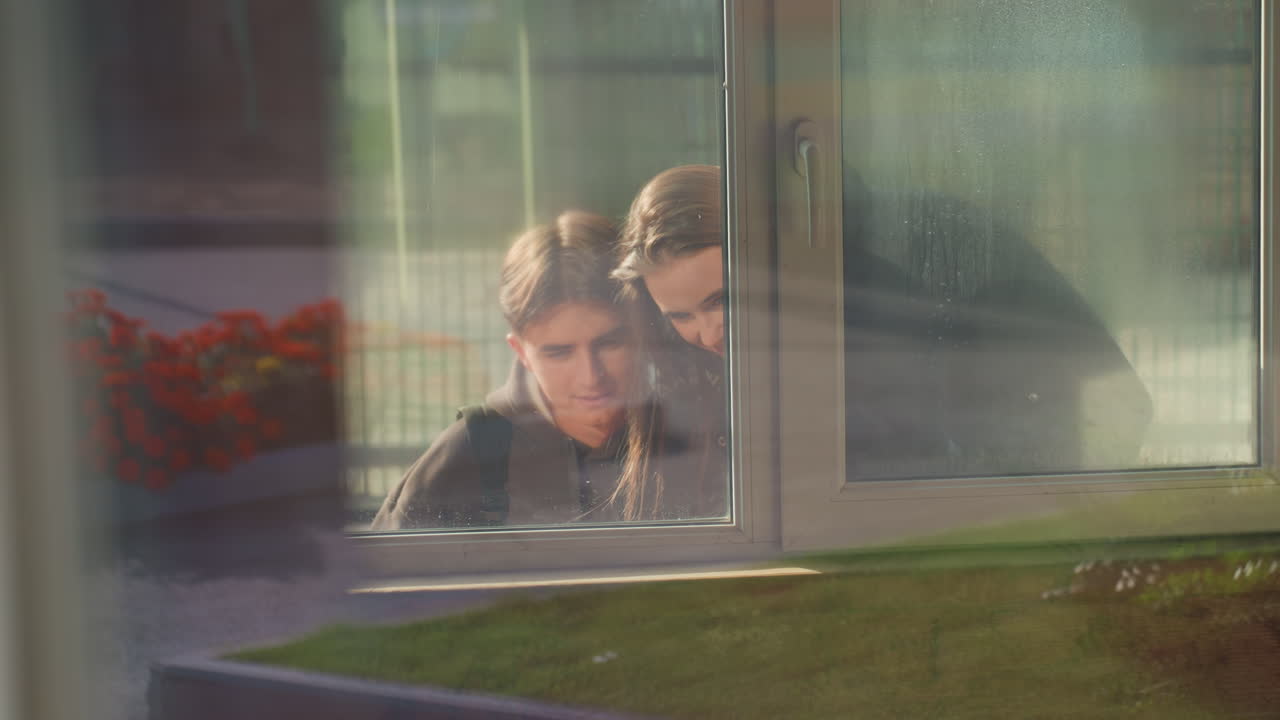 white teens peering through window, soft morning light and reflective glass reveal closeup faces, garden planter outside, hushed curiosity and quiet bond between siblings, cinematic composition