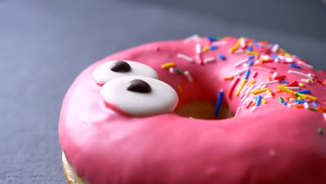 close-up upper-shot of tasty pink donut with multicolored chips and eyes spinning slowly on gray table background.