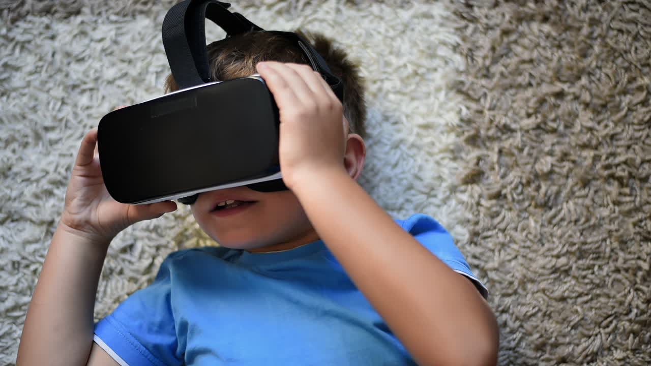 Blonde small boy lying on the carpet showing like and putting on VR headset