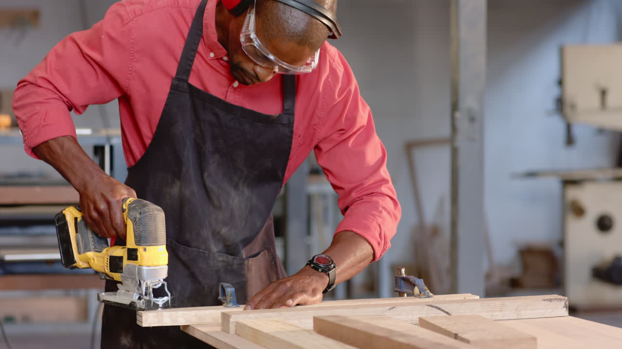 Operating African American man wearing red shirt and goggles cutting plank at workshop, with jigsaw