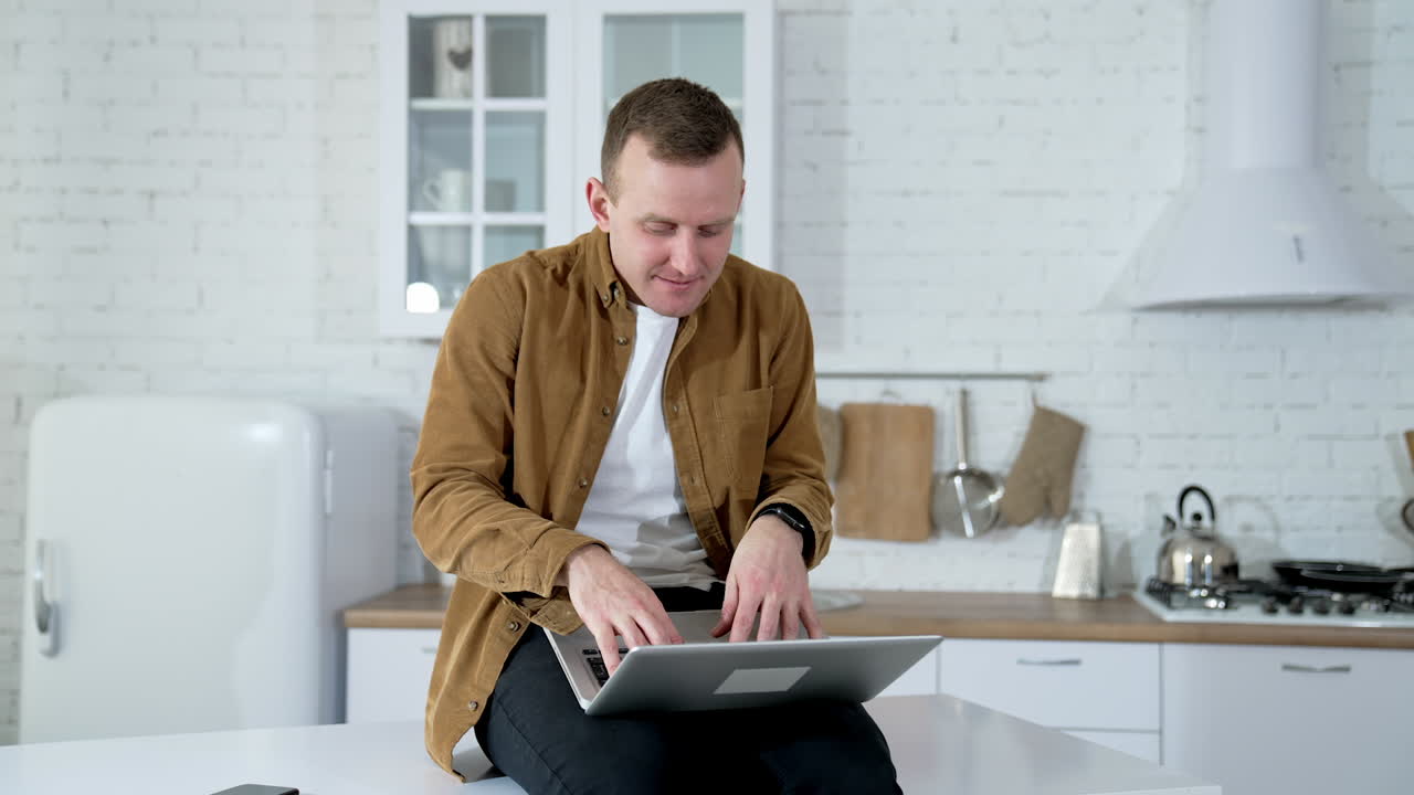 Funny freelancer typing on a laptop at home. Young male with a laptop sitting on a kitchen table. Long quarantine period.