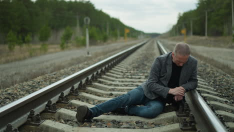 a man in a grey blazer and blue jeans lying on a railway track, looking contemplative and serious