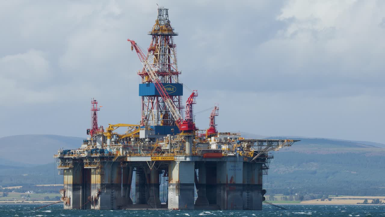 Stor offshore olieboreplatform stationær til søs, set fra kysten i dagslys med fjerne bakker og overskyet himmel, fanget i et stabilt vidvinkelbillede.