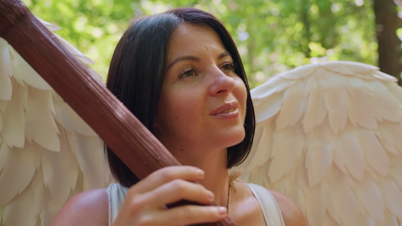 Angel woman with white wings holding wooden staff, eyes closed under soft forest sunlight, expressing harmony, spiritual peace, and gentle connection with nature surrounded by glowing green leaves
