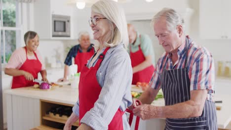 Feliz-Pareja-De-Ancianos-Caucásicos-Poniéndose-Delantales,-En-La-Cocina-Con-Diversos-Amigos,-Cámara-Lenta