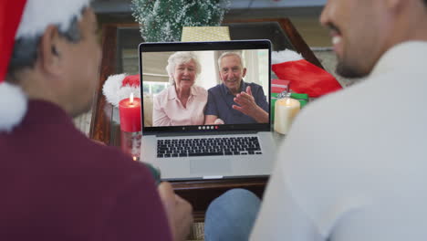 smiling biracial father with son using laptop for christmas video call with senior couple on screen