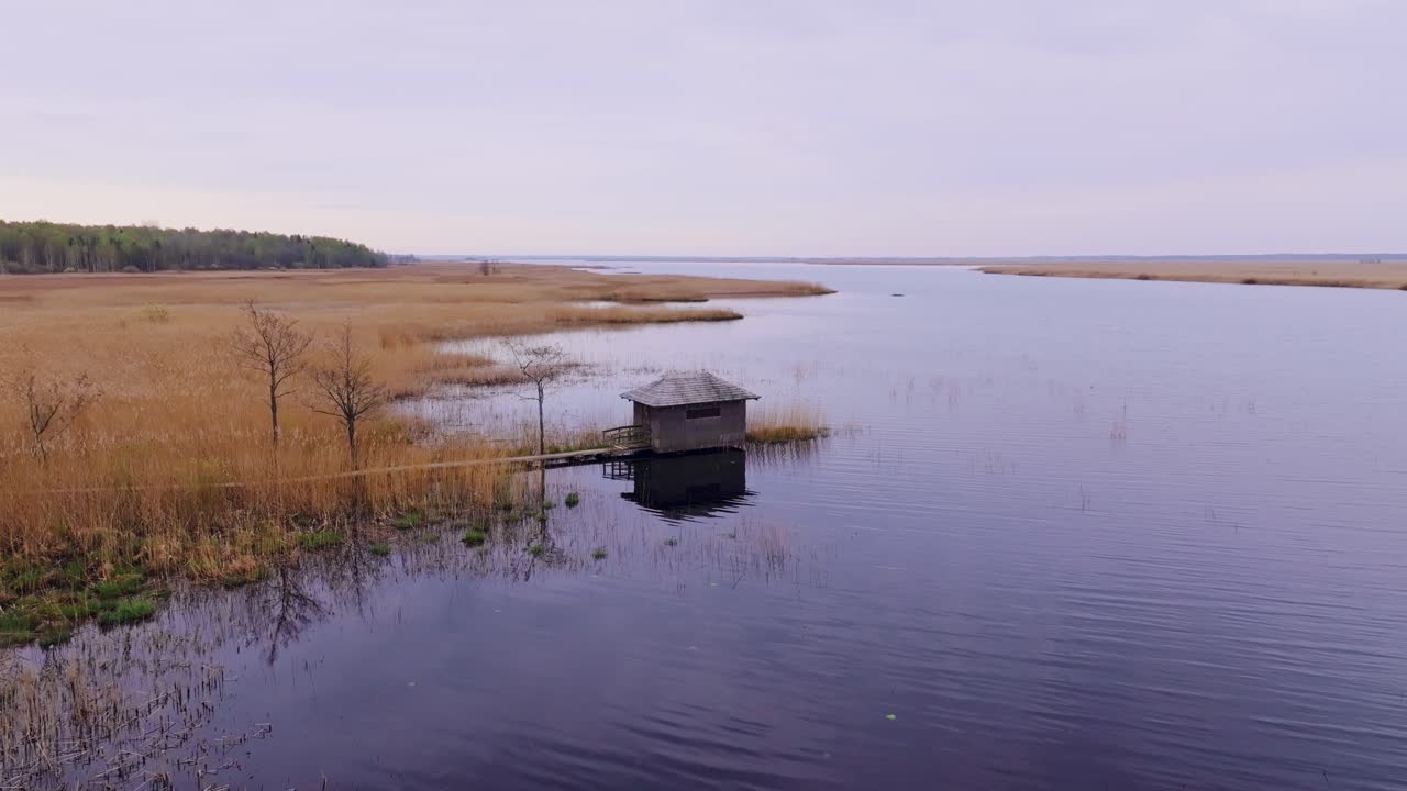 Drone slowly circles Kalnišķi birdwatching shelter over calm water, Lake Pape