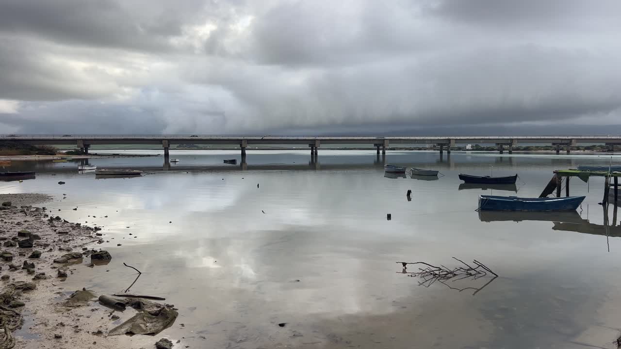 Barbate bridge reflecting on the tranquil lagoon, with boats gently floating under a moody, overcast sky in Andalusia, Spain, creating a serene and peaceful landscape