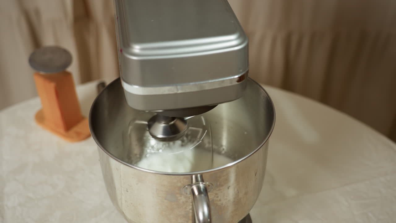 Close up of electric stand mixer whipping white ingredients inside stainless steel bowl on kitchen table, spinning rapidly beside wooden kitchen tool, with soft beige curtain background