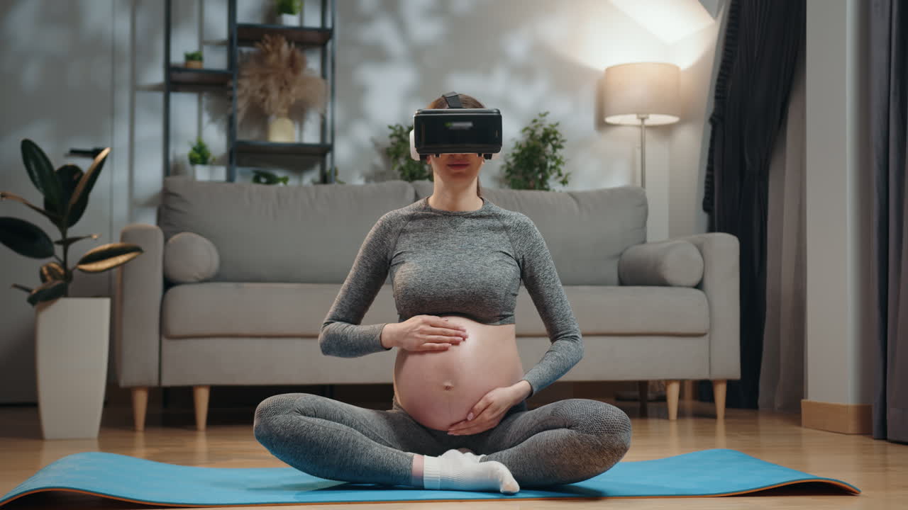 Pregnant woman wearing vr headset meditating in lotus pose on yoga mat in living room holding belly