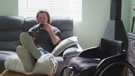 hombre discapacitado caucásico feliz sentado en el sofá en la sala de estar usando un teléfono inteligente y bebiendo café