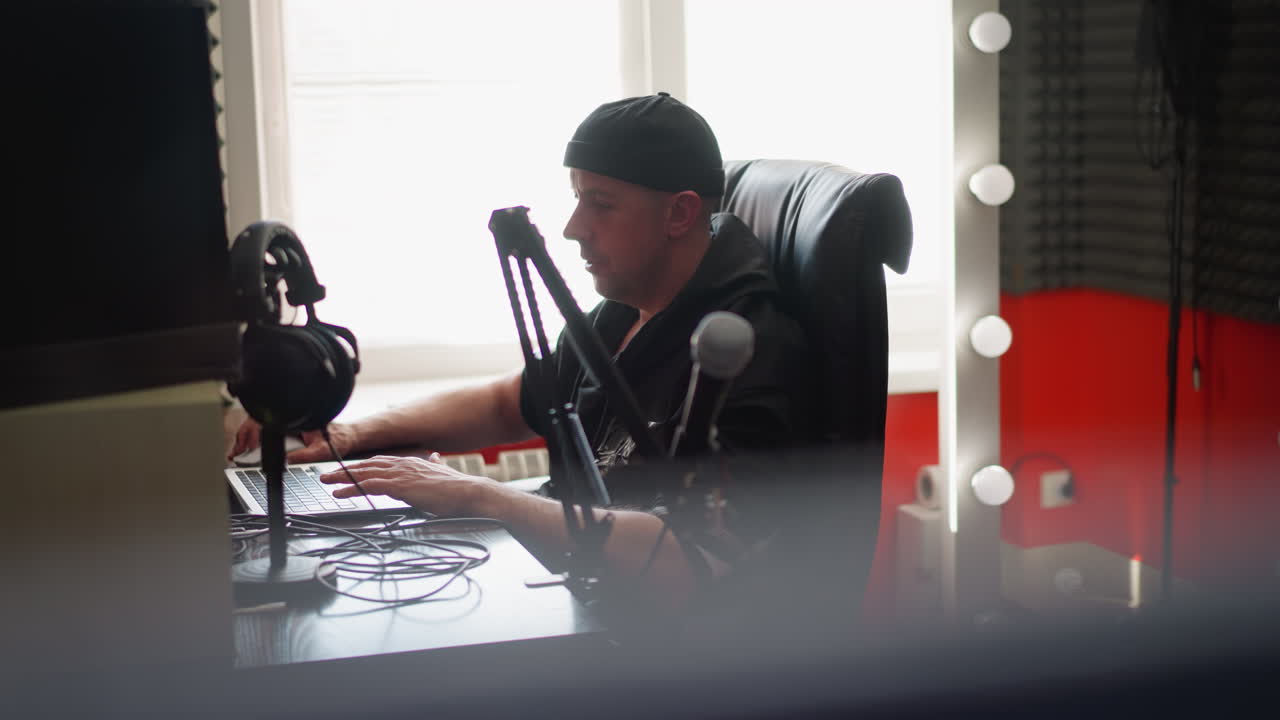 un hombre, probablemente un productor de música o podcaster, está sentado frente a una computadora en una habitación de estudio, con ropa negra y una gorra. el estudio está equipado con micrófonos y iluminación ambiental