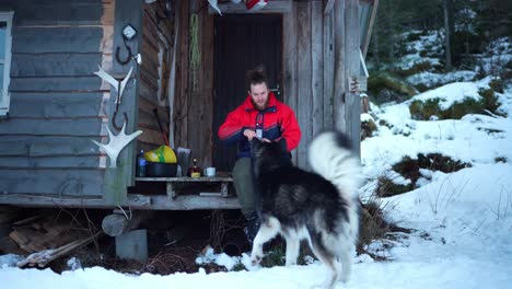 man sitting outside the wooden cabin eating with his dog during winter