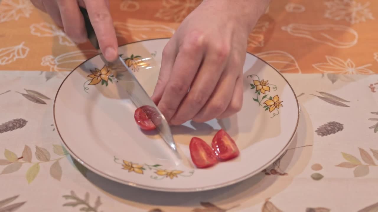 Close-up footage of fresh cocktail tomatoes being sliced with a knife on a decorative plate. Food preparation video with hands only