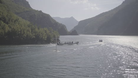 A-Ferry-trip-in-Albania-from-Fierze-to-Koman-over-the-lake-with-a-majestic-nature-around-on-a-sunny-day-with-clouds-looking-over-the-river-while-two-boats-speeding-and-passing-by-going-forward-LOG