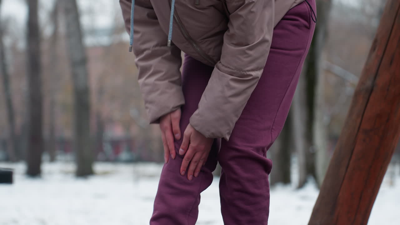 겨울 옷을 입은 여성이 구부러지고 무 통증이 느지고 배경에 벌거벗은 나무와 무을 문지르고, 바닥에 눈이 쌓여 있고, 추운 야외 겨울 환경에서 불편함을 나타니다.