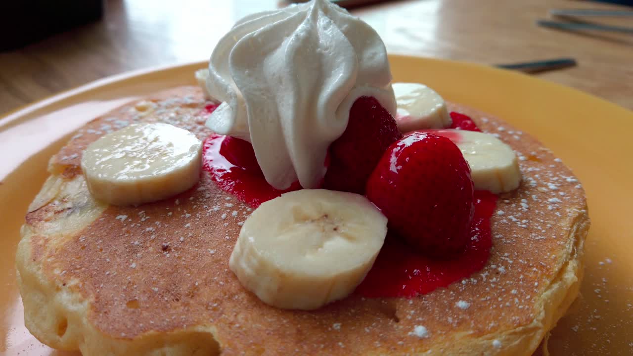 Close-up zoom out of pancakes topped with banana, strawberries and whipped cream.