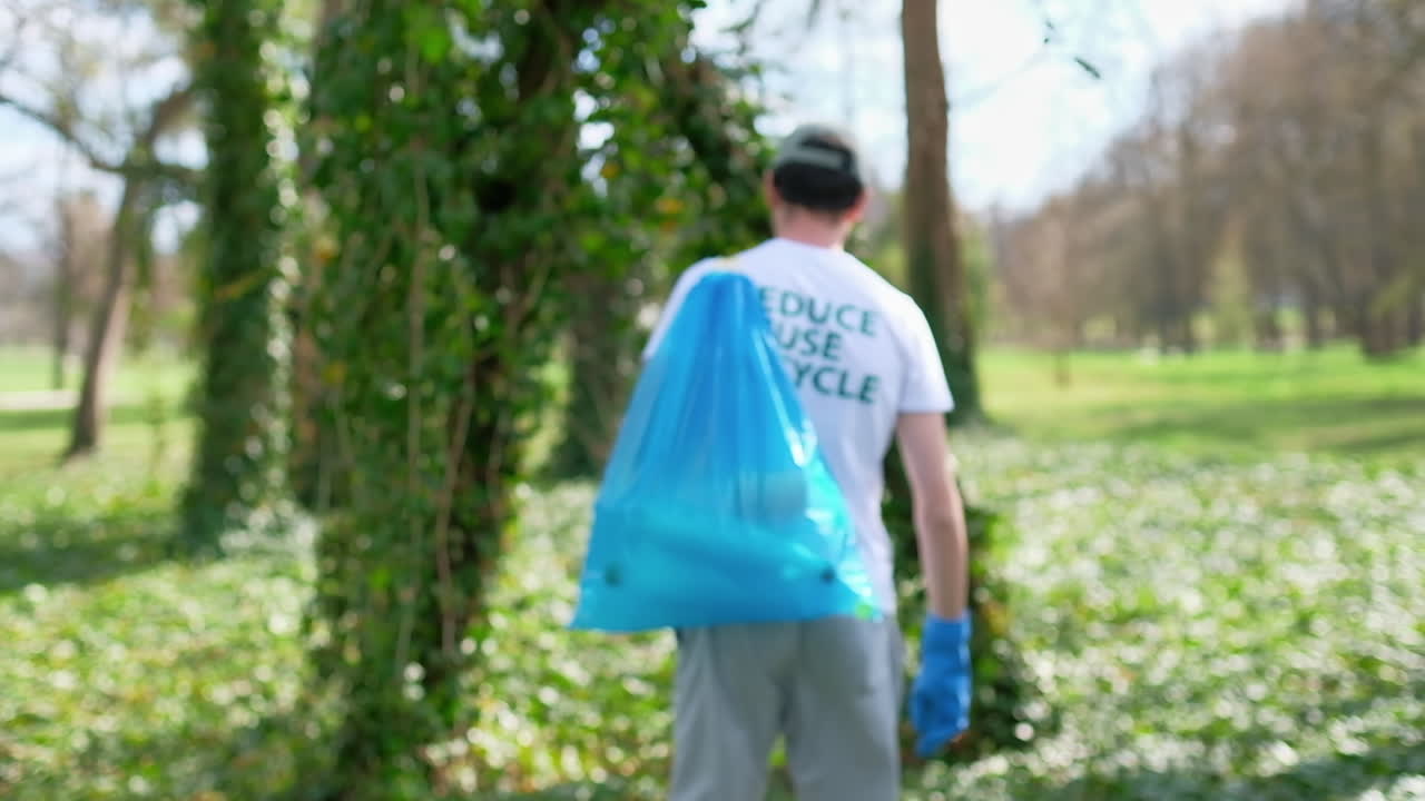 Man at plastic garbage collecting in a polluted park. Rubber gloves, carrying a bag with plastic bottles. Eco inscription on the T-shirt. Slow motion