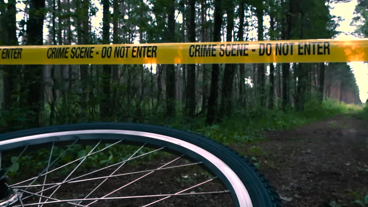 Low angle footage of fallen bicycle lying sideways on wooded forest road, wheel spinning slowly in eerie forest. Yellow crime scene tape across trail marking incident investigation site during evening