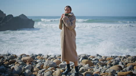 young woman standing shore in cozy coat. foaming ocean waves crash rocky coast