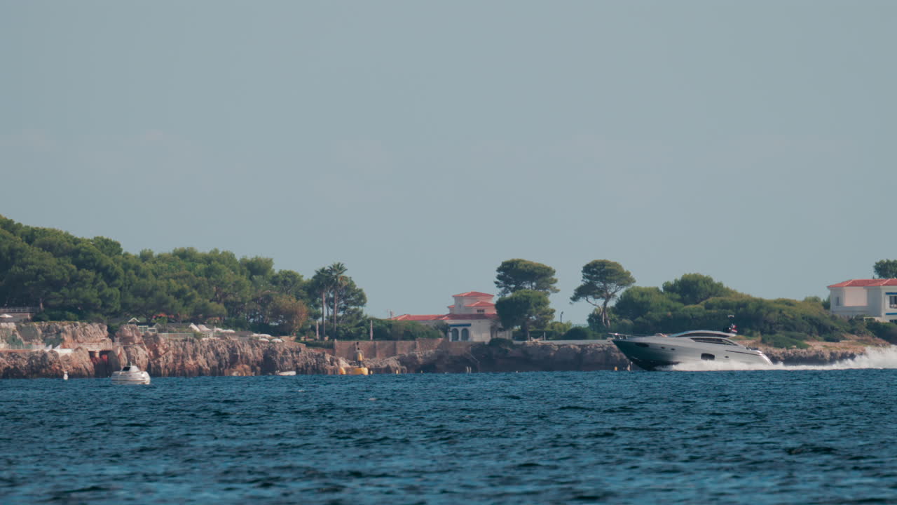 A fast moving yacht glides past a luxury coastal villa surrounded by trees and clear blue water