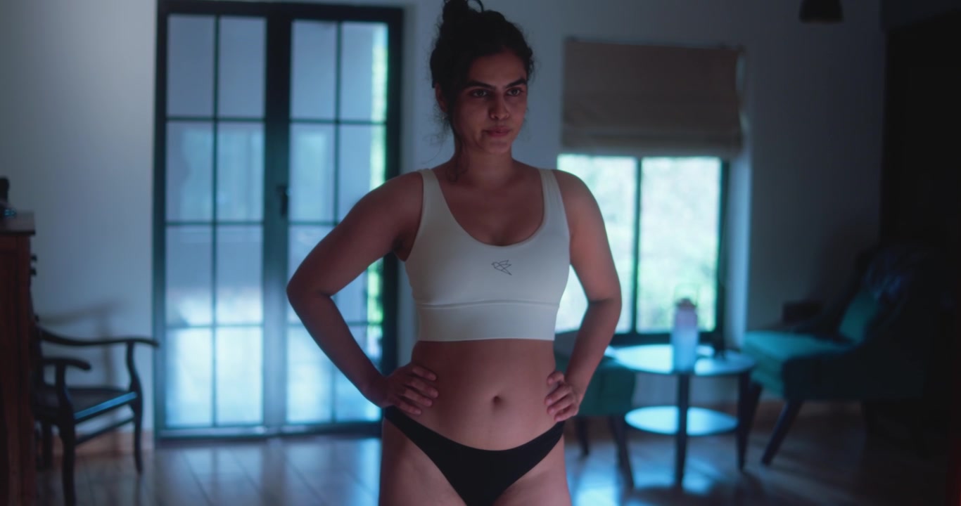 Giovane donna in reggiseno sportivo in piedi al chiuso dopo l'esercizio, che riprende fiato con le mani sui fianchi durante un momento di recupero a casa.