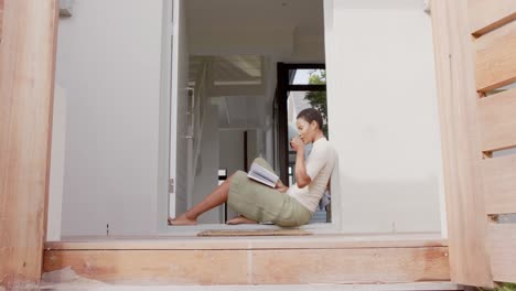 Mujer-Afroamericana-Sentada-En-La-Puerta,-Leyendo-Un-Libro-Y-Bebiendo-Té,-Cámara-Lenta