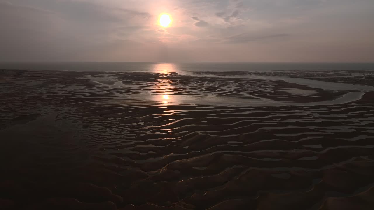 Sunset on sand patterns and pool reflections with sun low on horizon at low tide on coastline. Aerial pan. Half speed.