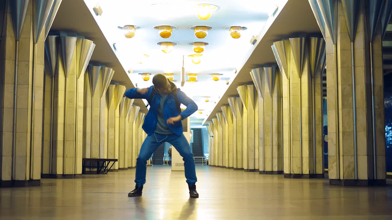 Gözlüklü şık bir adam, metroda modaya uygun bir şekilde dans ediyor ve kollarını sallıyor. Genç adam kulaklıkla müzik dinliyor ve istasyonda zıplıyor.