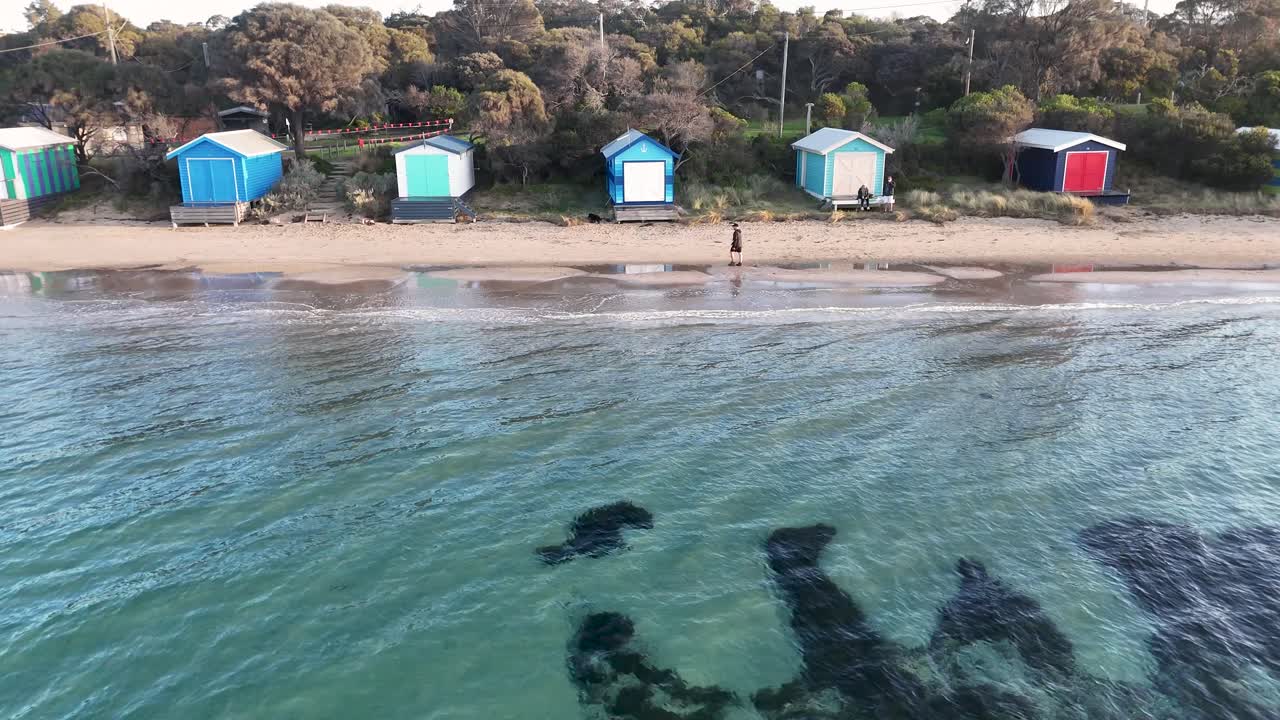 Drone glides above clear water, revealing vibrant beach huts, sandy shore, and tranquil coastal scenery